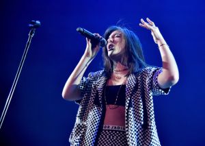 Lorde singing with her eyes closed wearing a checkered outfit holding the mic with one hand and raising her other hand up.