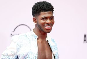 Lil Nas X smiling wearing an open white and blue graphic jacket.
