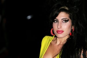 Amy Winehouse looking right wearing red hoop earrings and a bright yellow top.
