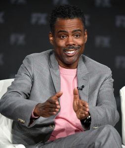 Chris Rock wearing a grey suit with a pink shirt.