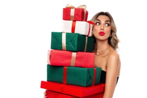 Holiday shopping season. Young pretty woman holding huge stack of many Xmas or New Year wrapped gifts against white wall, excited female with red lipstick in evening dress receiving Christmas presents