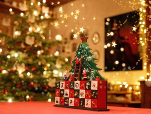 painted advent calendar made of wood with stylised Christmas Tree, tiny figures and drawers standing on a table in the middle of a Christmassy decorated and illuminated family room