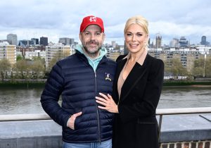Jason Sudeikis smiling wearing a blue down jacket with his hand in his pocket smiling wearing a red baseball cap and Hannah Waddingham wearing a black blazer with her hand on his chest during the photocall for "Ted Lasso" Season 3