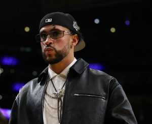 Bad Bunny walks courtside during the game between the Golden State Warriors and the Los Angeles Lakers wearing a leather jacket and white collared shirt with a backwards baseball cap.