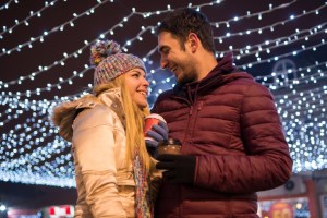 Close-up front view image of a young cheerful couple in winter clothing having warm drink at night during winter holidays. Luminous decoration background