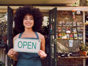 Smile of a retail store entrepreneur with welcome shopping board and business owner with plant products