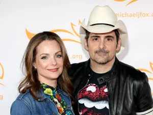 Brad Paisley is wearing a black jacket and cowboy hat and his wife Kimberly wearing a colorful dress and denim jacket