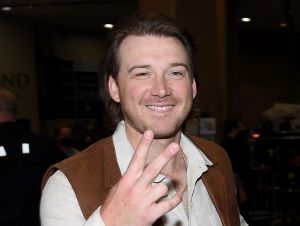 Morgan wallen holding his fingers up in a beige shirt and brown vest