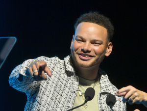 Kane Brown on stage pointing wearing a green and beige flannel and a tan t-shirt
