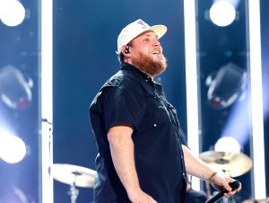 Luke Combs on stage smiling wearing a black shirt and white ball cap