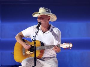 Kenny Chesney on stage playing guitar in a white polo shirt and cowboy hat
