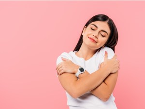 smiling pretty brunette girl hugging herself with closed eyes isolated on pink, single, self love concept