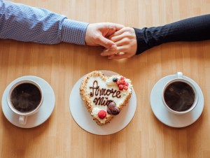 Flat lay, top view of man and woman holding hands while drinking coffee with cream cake with the Italian language text "Amore mio" ("My Love"), love language concept