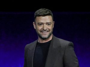 Justin Timberlake speaks onstage as he promotes the upcoming film "Trolls Band Together" during the Universal Pictures and Focus Features presentation during CinemaCon smiling wearing a grey blazer.