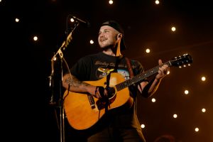 Zach Bryan wearing black playing a guitar on stage