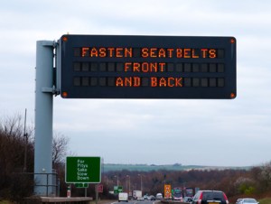 A large illuminated over-road traffic sign gives a message warning drivers and passengers to wear their seat belts. A further green road sign implores them to slow down.