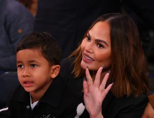 Chrissy Teigen and her son watch the game between the New York Knicks and Milwaukee Bucks at Madison Square Garden facing left putting her right hand up.