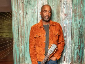 Darius Rucker posing in a brown jacket and green shirt holding a guitar at his feet.