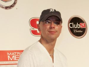 Kenny Chesney is posing in a blue ball cap, glasses, and a white shirt.