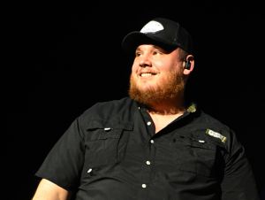 Luke Combs on stage wearing a black button-down shirt and a black ball cap.