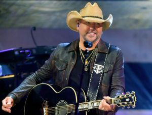 Jason Aldean performing on stage with a guitar wearing a black leather jacket and a cowboy hat.