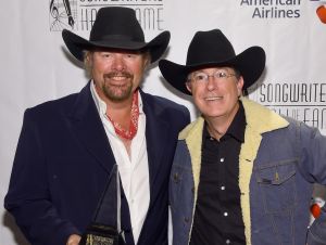 Toby Keith in a black suit and black cowboy hat holding an award with Stephen Colbert in a cowboy hat and denim coat.