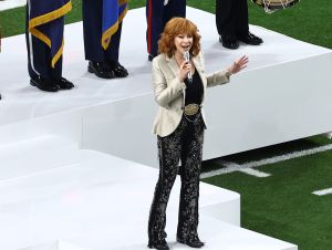 Reba McEntire is wearing black pants and a tan blazer, singing the National Anthem at the Super Bowl.