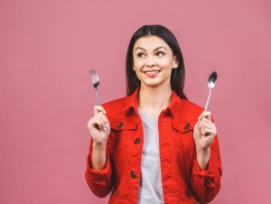 Time for dinner! Portrait of young beautiful woman wearing red casual shirt, holding spoon and fork isolated over pink background.