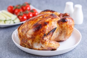 Chicken baked whole to a delicious crust. A traditional dish. In the background are cucumbers and cherry tomatoes on a white plate. Close-up, selective focus, gray background.