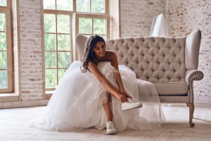 Attractive young woman in wedding dress putting on sports shoes and smiling while sitting on the sofa