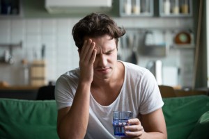Young man suffering from strong headache or migraine sitting with glass of water in the kitchen.