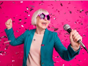 Photo of old mature stylish energetic woman singing in microphone, wearing star shaped spectacles standing in falling confetti isolated over pink vivid color background