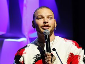 Kane Brown on stage holding a mic in a white, red and black patterned sweater.