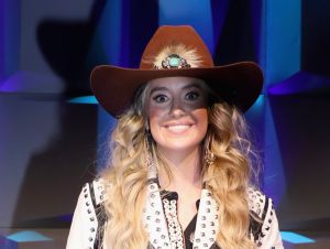 Lainey Wilson is posing in a brown cowboy hat and white leather studded jacket.