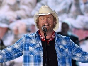 Toby Keith on stage in a blue plaid shirt, red bandana, and cowboy hat.