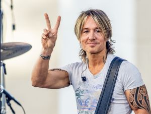 Keith Urban on stage in a gray t-shirt giving the peace sign.