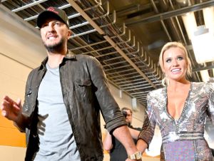 Luke Bryan, wearing a ball cap and an open black shirt, with his wife, Caroline, in a silver dress, walking backstage.