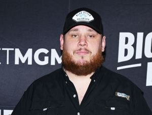 Luke Combs posing in a black ball cap and black button-down shirt.