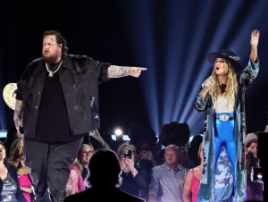 Jelly Roll wearing black on stage with Lainey Wilson in blue pants and a jacket and hat.
