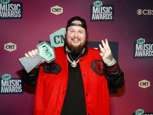 Jelly Roll on the 2023 CMT Music Awards red carpet wearing a red jacket and holding an award.