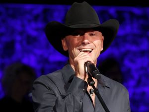 Kenny Chesney is smiling on stage, wearing a black shirt and black cowboy hat.