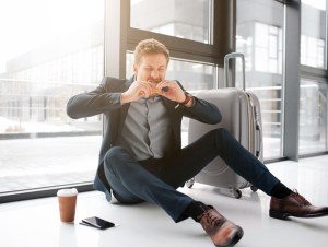 Hungry young man devour burger. He sit on floor in airport. Phone stand on floor with cup of coffee. Suitcase is there as well, airport fast food concept.