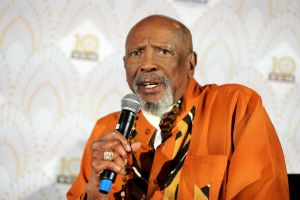 Louis Gossett Jr. on stage speaking into a microphone. he's wearing an orange shirt.