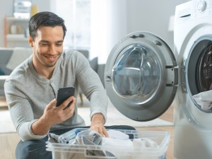 miling Young Man in Grey Jeans and Coat Sits in Front of a Washing Machine and Uses His Smartphone, man doing chores with phone in hand.