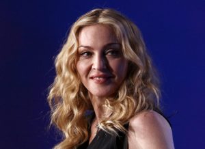 Madonna looks on on during a press conference for the Bridgestone Super Bowl XLVI halftime show smiling looking right wearing a black tank top with medium length golden wavy hair.