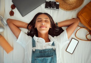 Top view, relax and woman sleeping on bed at hotel on vacation, holiday or trip. Phone mockup, travel dream and happy female in bedroom at motel, resort or lodge with camera, laptop and headphones