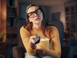 Woman sitting in the living room at home and watching horror movies on TV, she is holding the remote control and looking at camera, streaming netflix, rocku, concept.