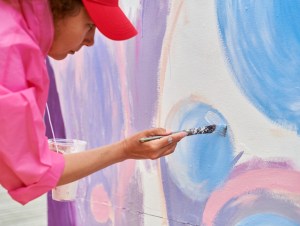 Female painter in cap draws picture with paintbrush on canvas for outdoor street exhibition, close up side view of female artist apply brushstrokes to canvas, symphony of art creativity