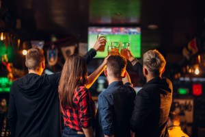A group of friends watch football on TV and raised their glasses with beer in a sports bar at night.