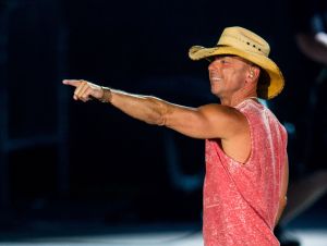 Kenny Chesney Is Deep In Tour Rehearsals - Kenny is on stage pointing, wearing a red sleeveless shirt and a cowboy hat.